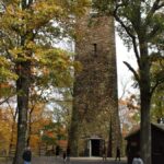 A large stone tower stands among tall trees in the fall while people walk around its base