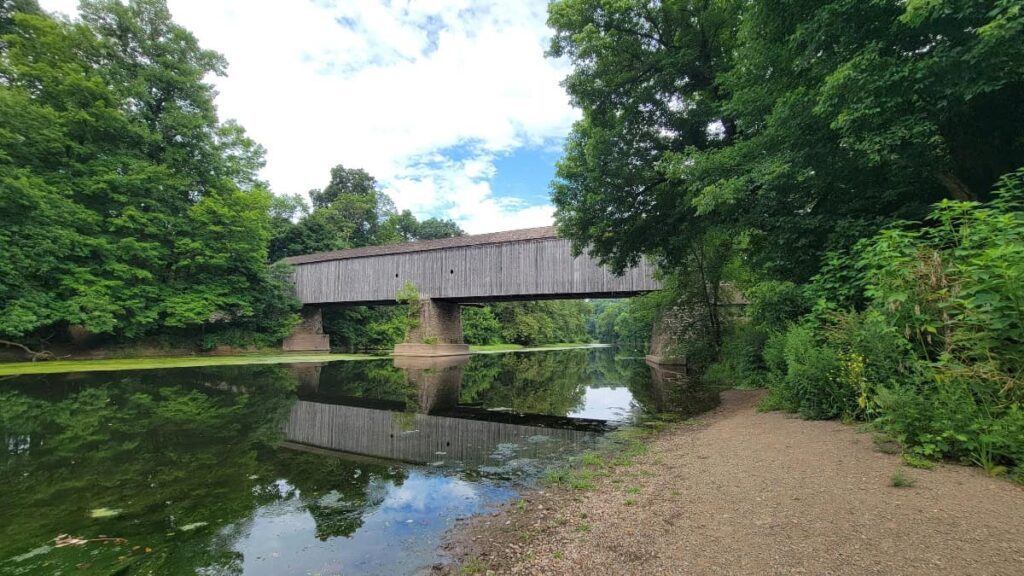 an aged gray wooden covered bridge spans over a creek with green trees on either side