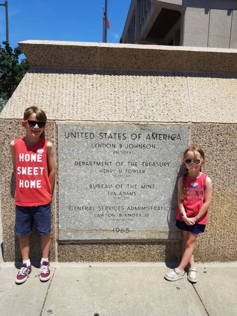 Two kids stand next to the engraved US Mint Sign outside of the mint in Philadelphia
