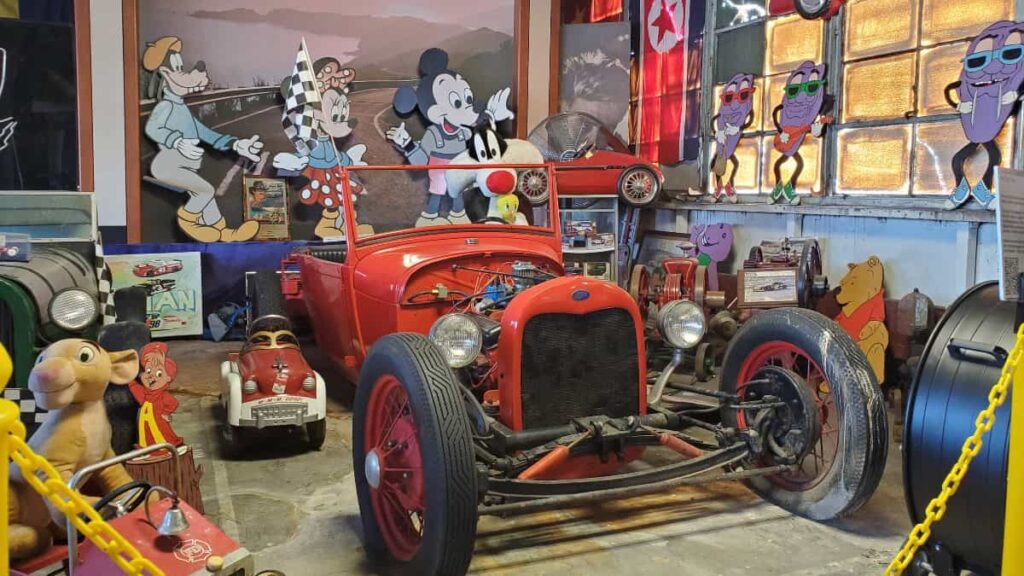 A red classic car sits in front of large signs depicting Mickey Mouse, Minnie Mouse, and other cartoon characters