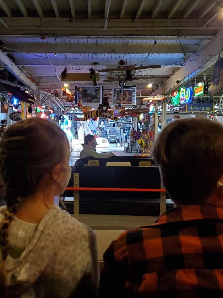 Two kids sit in front of viewer on a tram ride through a warehouse filled with memorabilia
